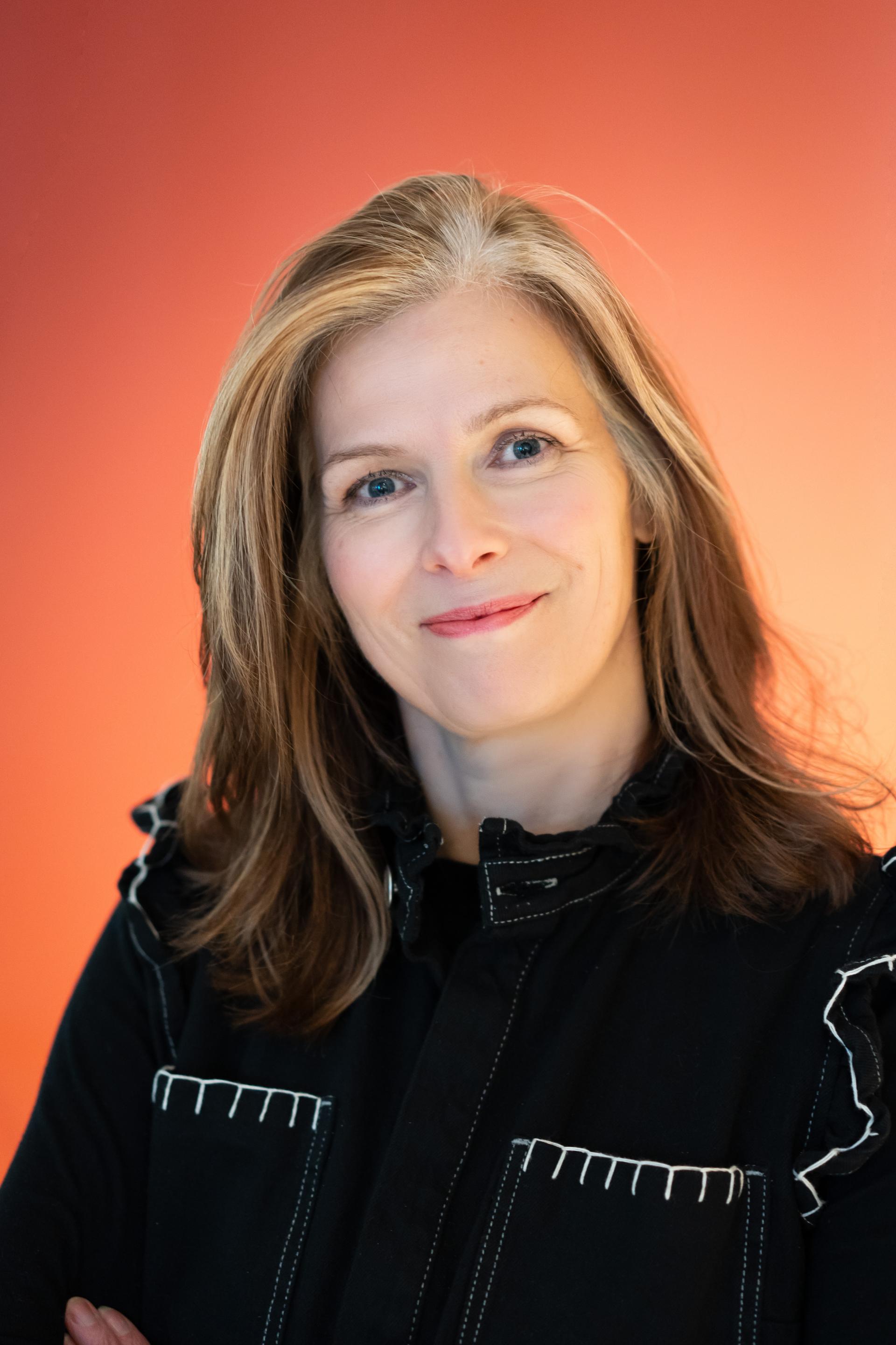 Business professional with long, brown hair smiling against a vibrant orange background, emphasizing workplace flexibility and leadership.