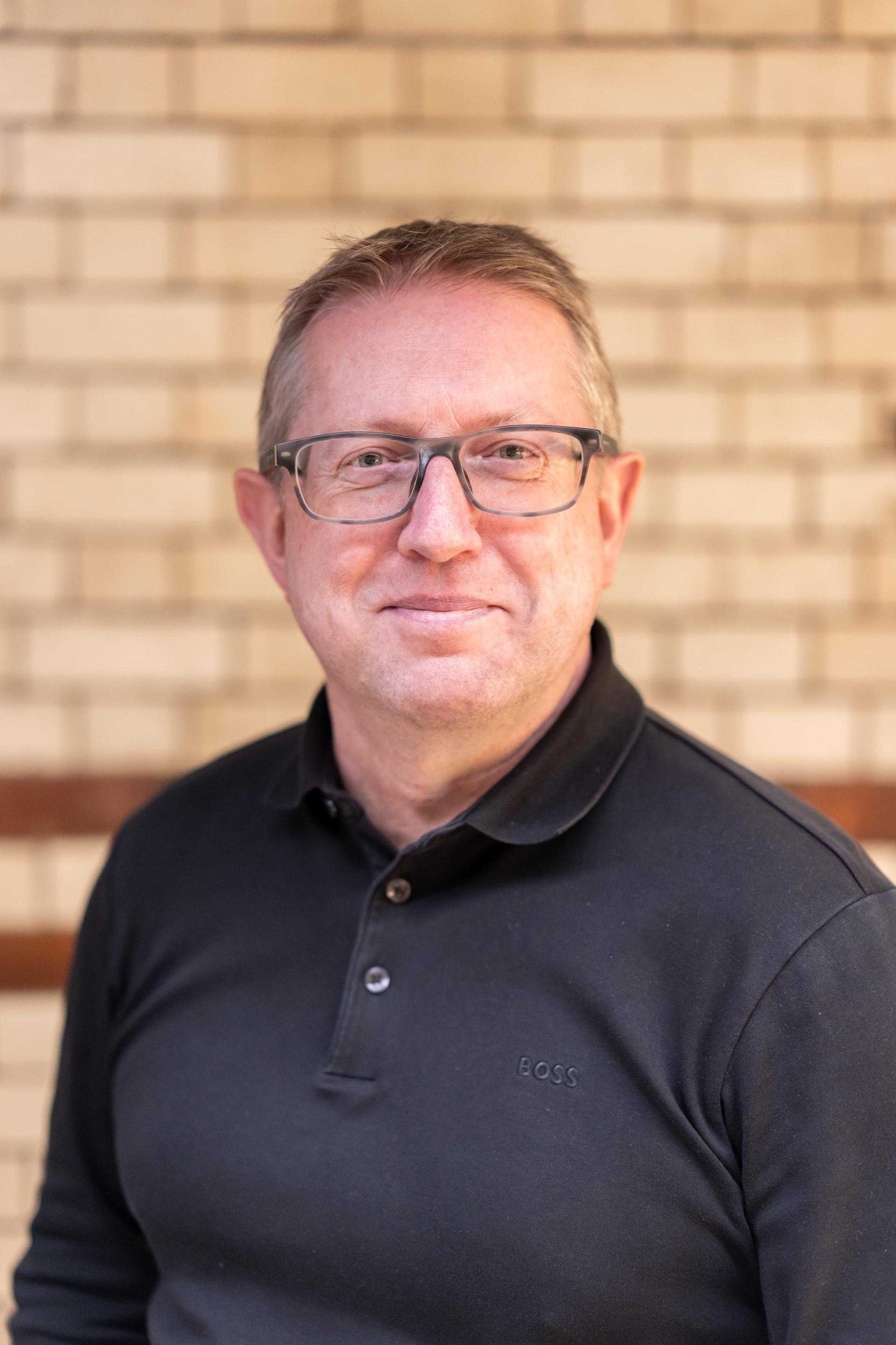 Professional looking man in a black polo shirt, smiling against a textured brick wall, representing EDI culture and engagement.