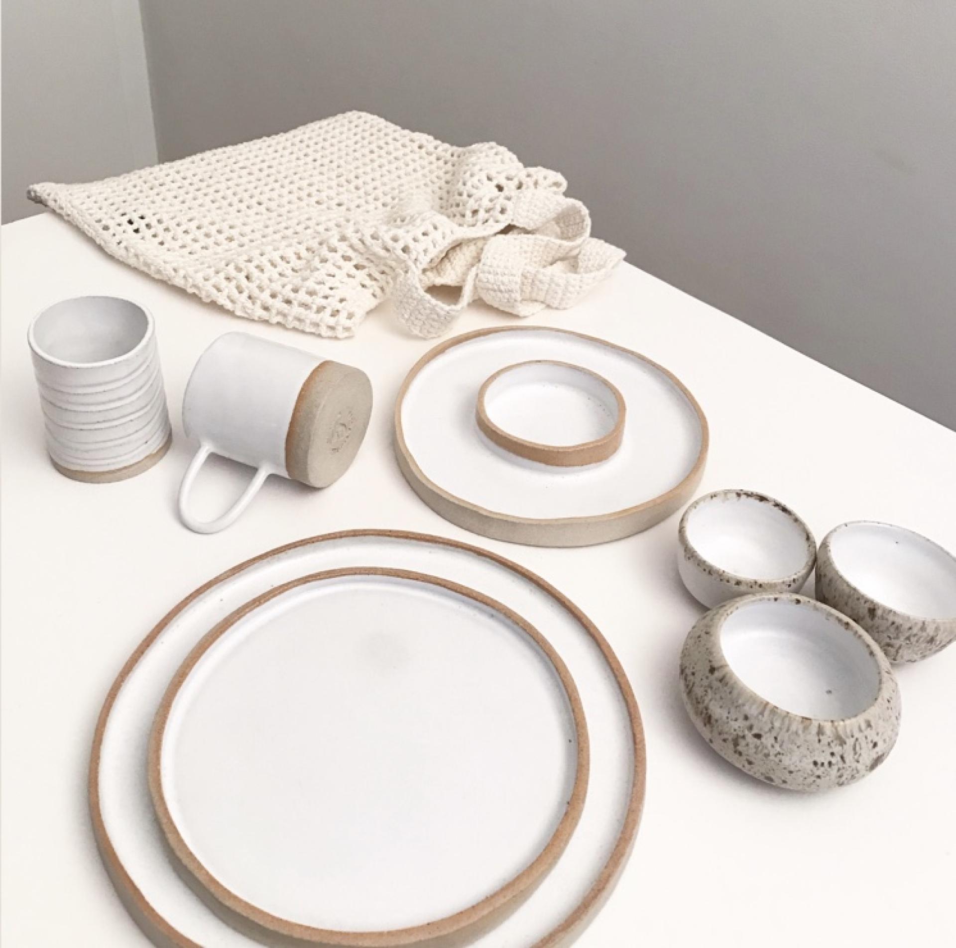 Handcrafted ceramics, including plates, bowls, and cups, displayed alongside a textured white cloth in a minimalist setting.