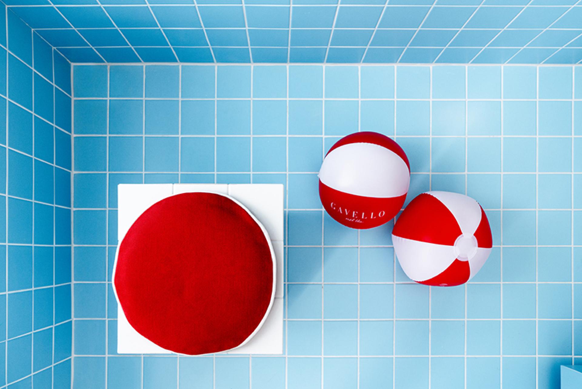 Vibrant blue swimming pool tiles featuring red beach balls and a round cushion, capturing a whimsical jewellery shop vibe.