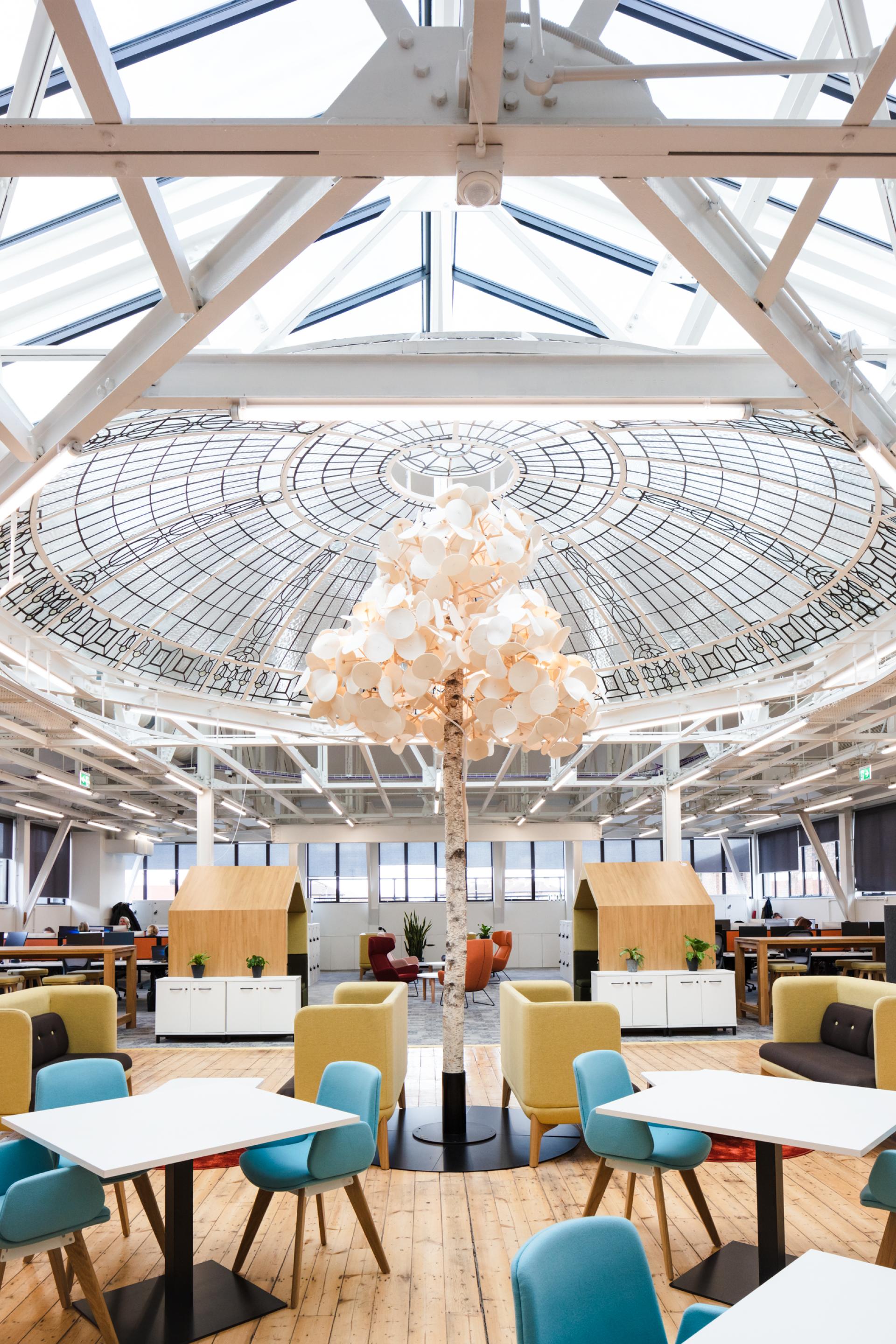 Modern workspace at Queen Mary University featuring collaborative seating, an artistic tree centerpiece, and abundant natural light.