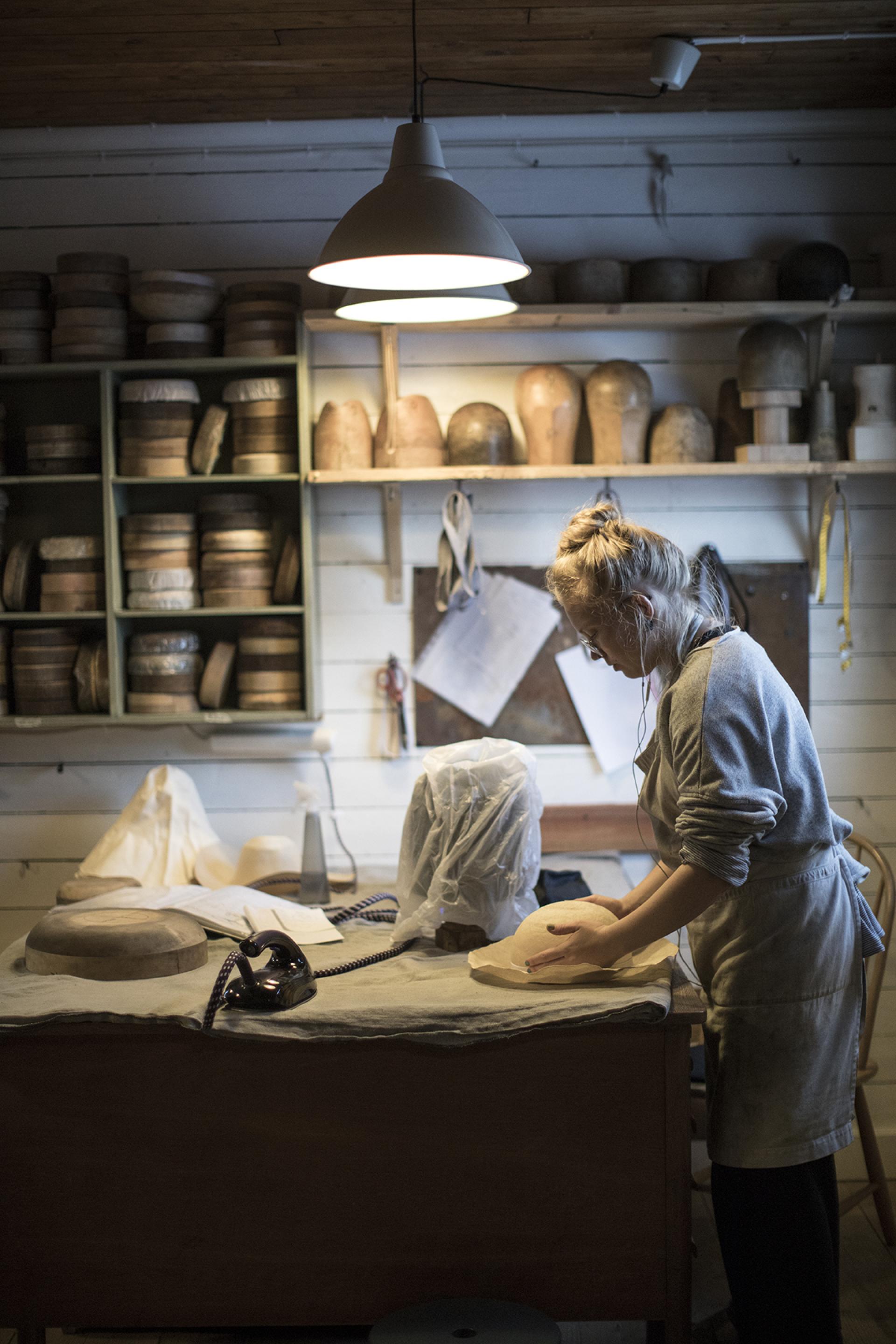 Craftsperson working in Horisaki's hatmaking workshop, with wooden molds and fabric on the table in Småland's forest setting.
