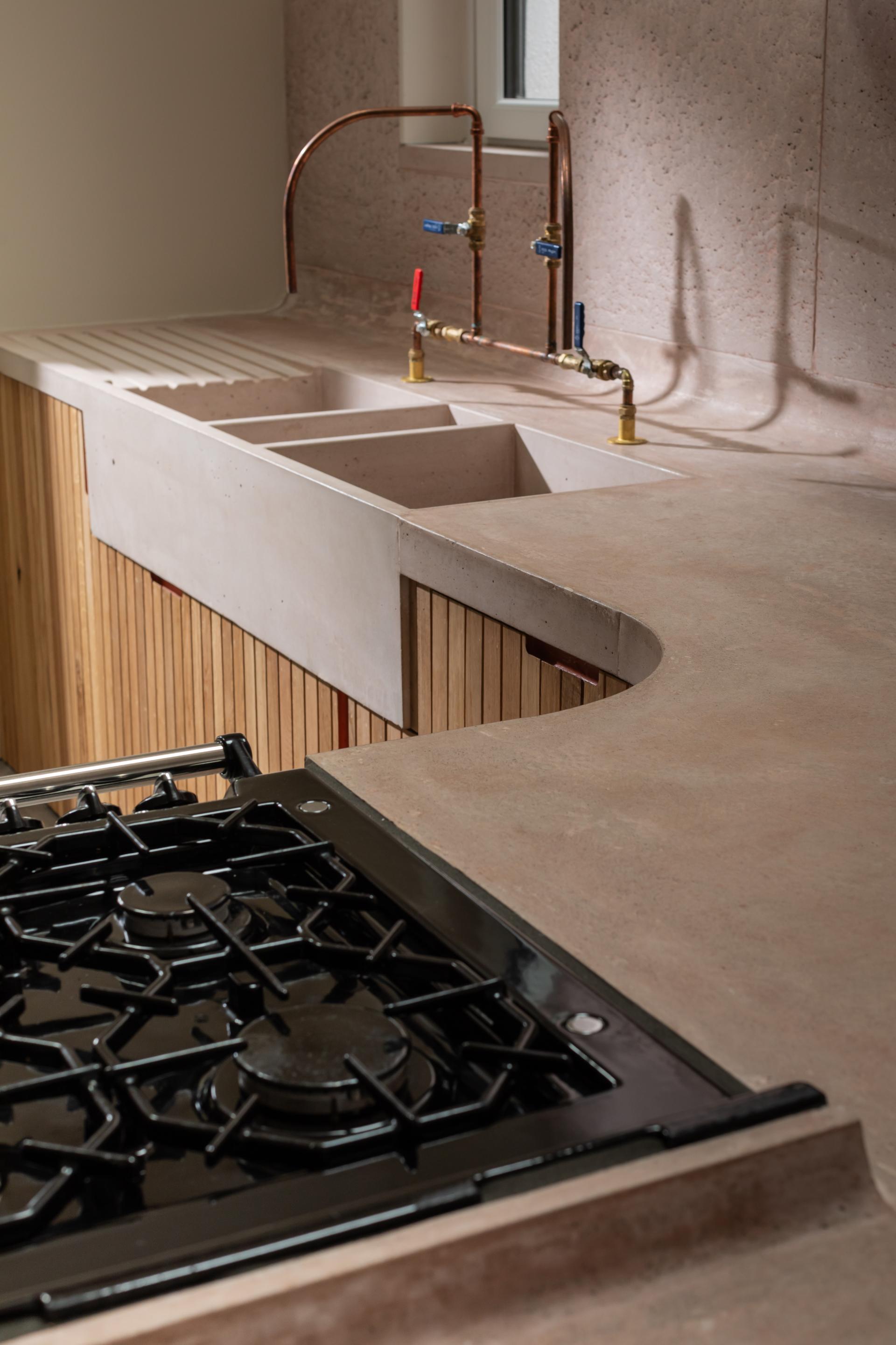 Residential kitchen featuring curved pink concrete countertops, a sleek black gas stove, and warm Scottish Oak cabinetry.