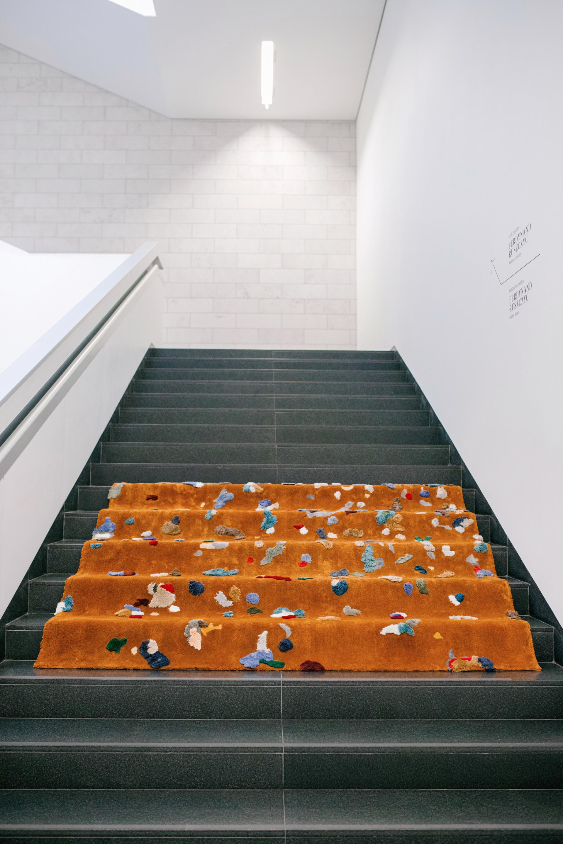 A colorful chaos rug by Audronė Drungilaitė elegantly covers a staircase, adding artistic flair to the space.