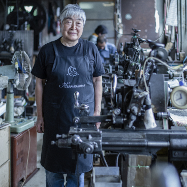 Craftsman in Tsubame-Sanjo workshop stands proudly among tools, highlighting Japan's rich metal craftsmanship in Biology of Metal exhibition.