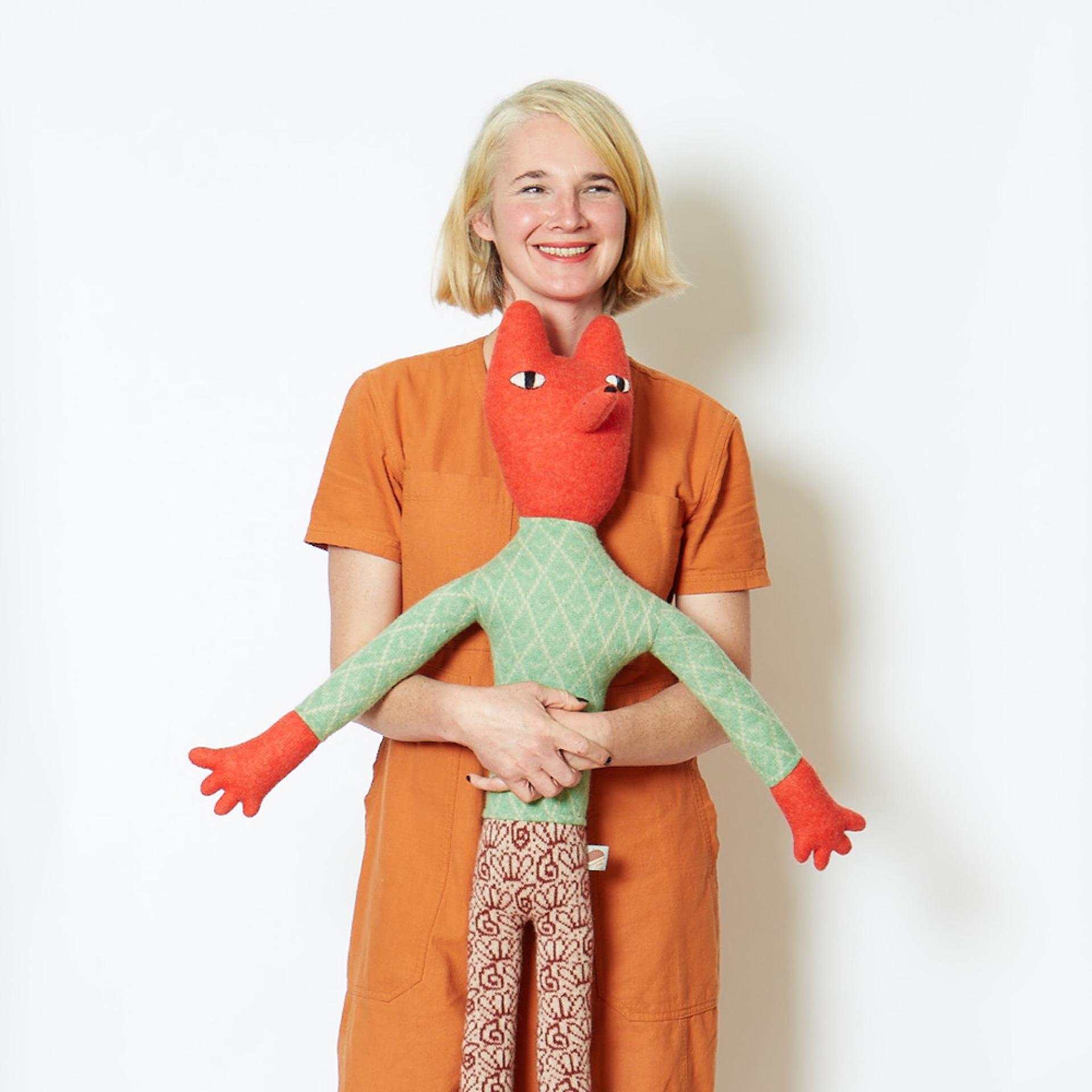 A smiling woman in an orange outfit holds a colorful textile puppet, showcasing creativity and design at Dundee Design Festival 2024.