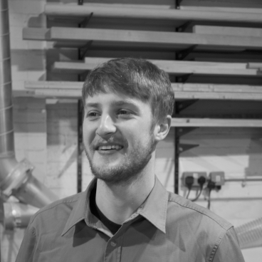 Jesse Cracknell, the maker in residence, smiling in a workshop setting with wood and tools in the background.