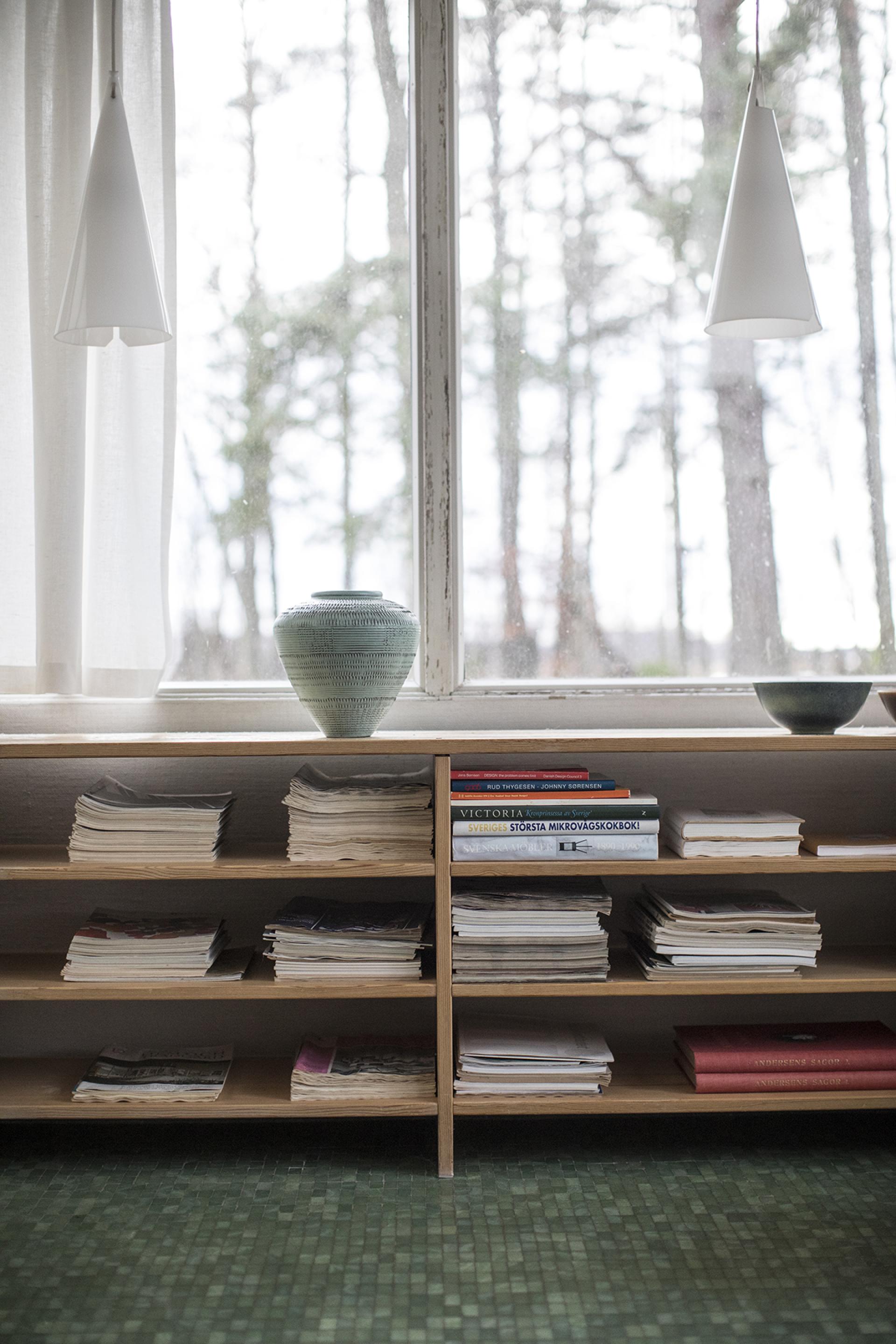 Södra Kull interior featuring shelves with books, magazines, and a decorative vase by large windows surrounded by nature.
