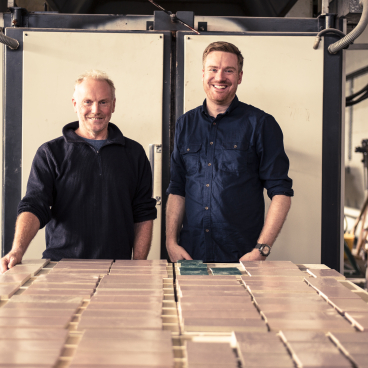 Two men standing in a workshop, showcasing sustainable surface materials against a backdrop of equipment and materials.