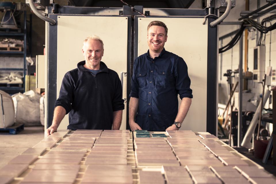 Two men standing in a workshop, showcasing sustainable surface materials against a backdrop of equipment and materials.