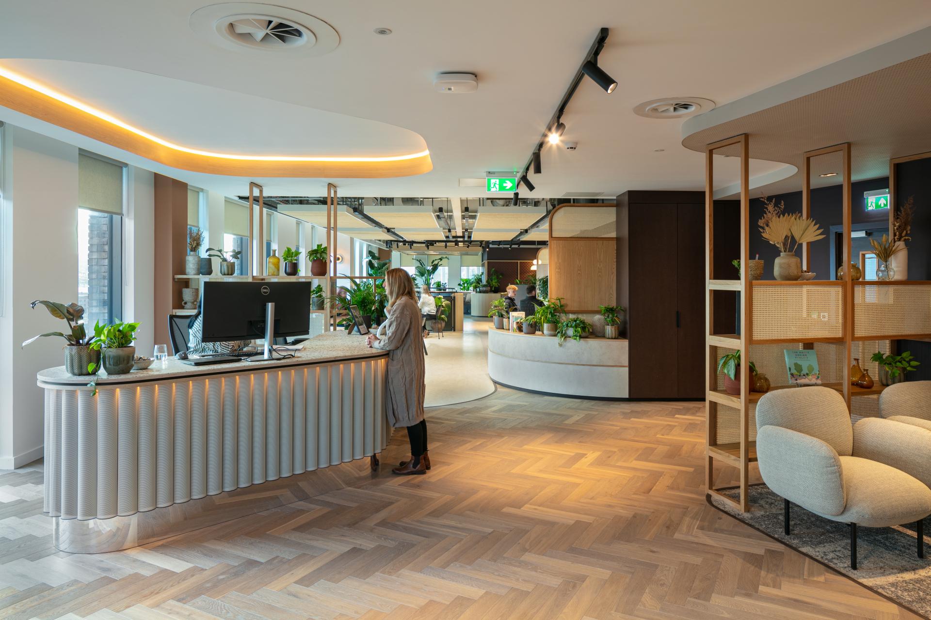 Modern office reception area featuring plants, wood accents, and a woman at the front desk.