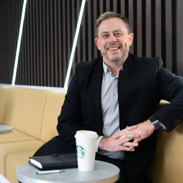 Lee Birchall smiles while seated at a table with a Starbucks cup, discussing team dynamics at DV8 Designs.