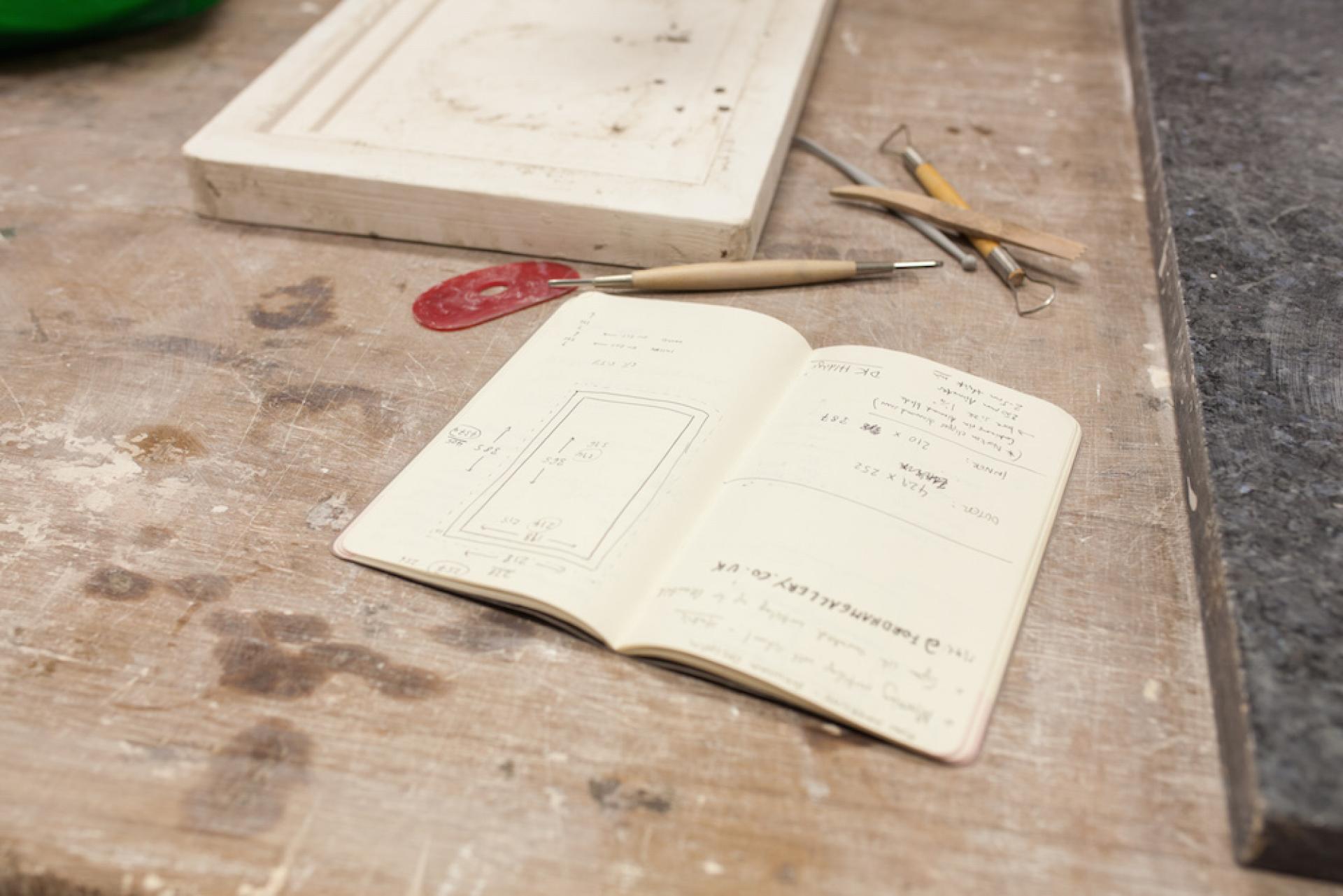Open notebook with clay design sketches and measurements on a workbench, surrounded by ceramic tools in a studio setting.