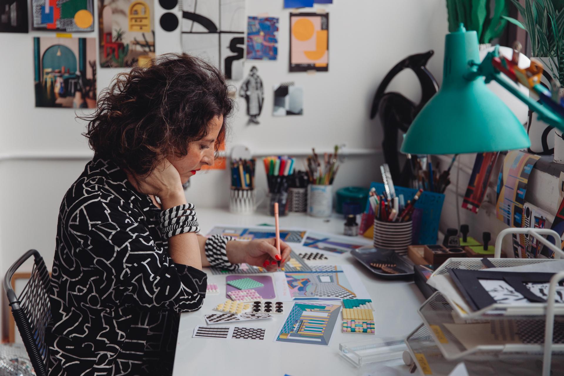 Camille Walala sketches design concepts at her desk, surrounded by vibrant materials and art in her creative studio.
