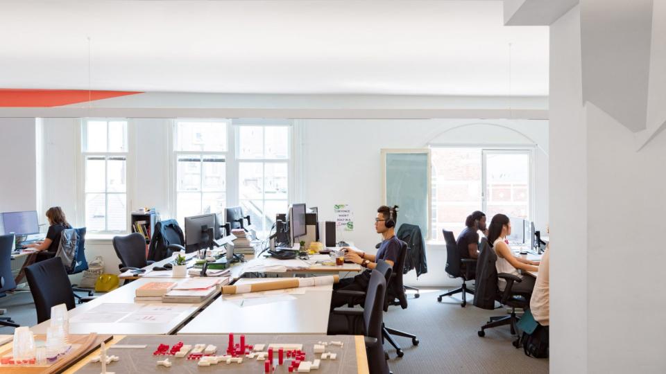 Architects working in a collaborative studio environment, featuring computers, models, and large windows for natural light.
