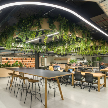 Modern collaborative workspace at Glasshouse, featuring greenery, communal tables, and individuals engaging with technology.