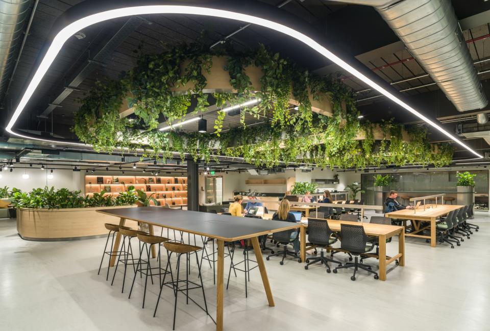 Modern collaborative workspace at Glasshouse, featuring greenery, communal tables, and individuals engaging with technology.