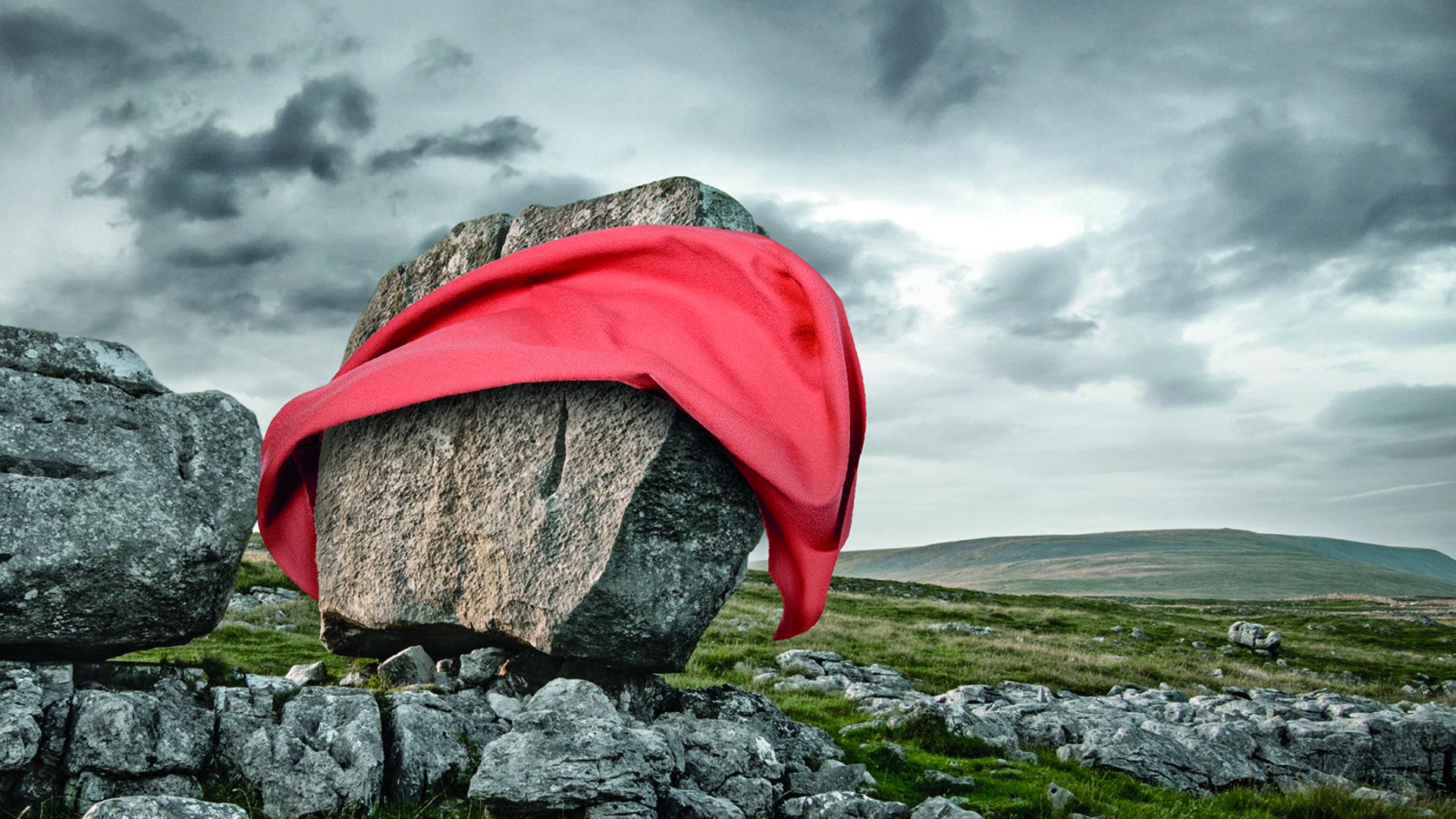 A large rock draped with vibrant red fabric against a cloudy sky in a natural landscape, symbolizing sustainable design.