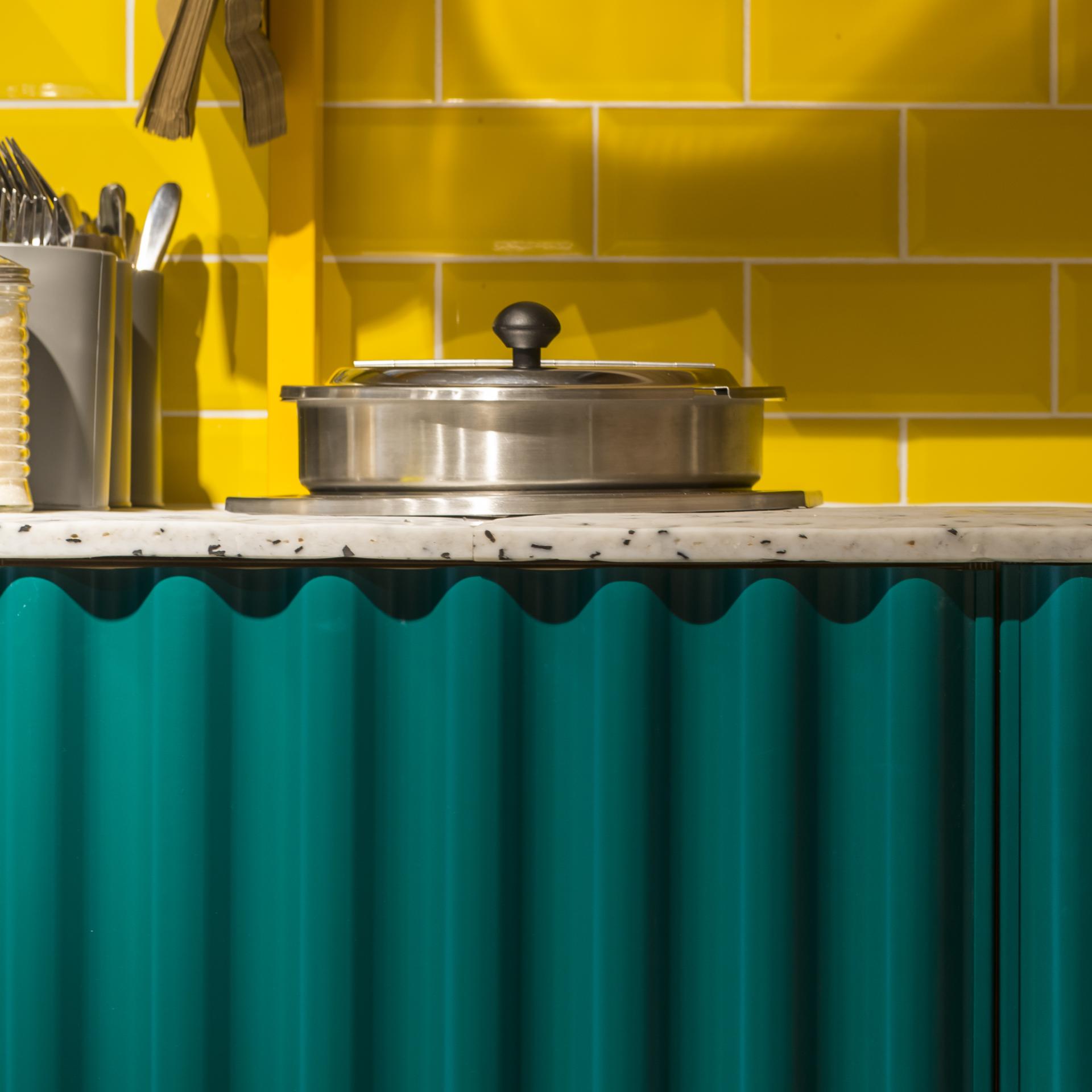 Colorful café-deli countertop showcasing a stainless steel pot, vibrant yellow tiles, and teal decorative elements.