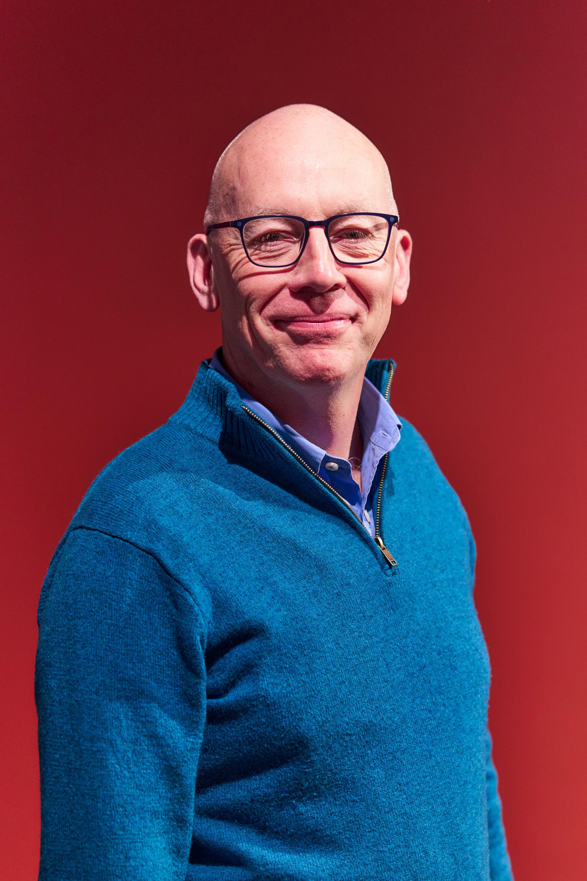 Smiling man in a blue sweater poses against a vibrant red background during the Glasgow Property Outlook seminar.