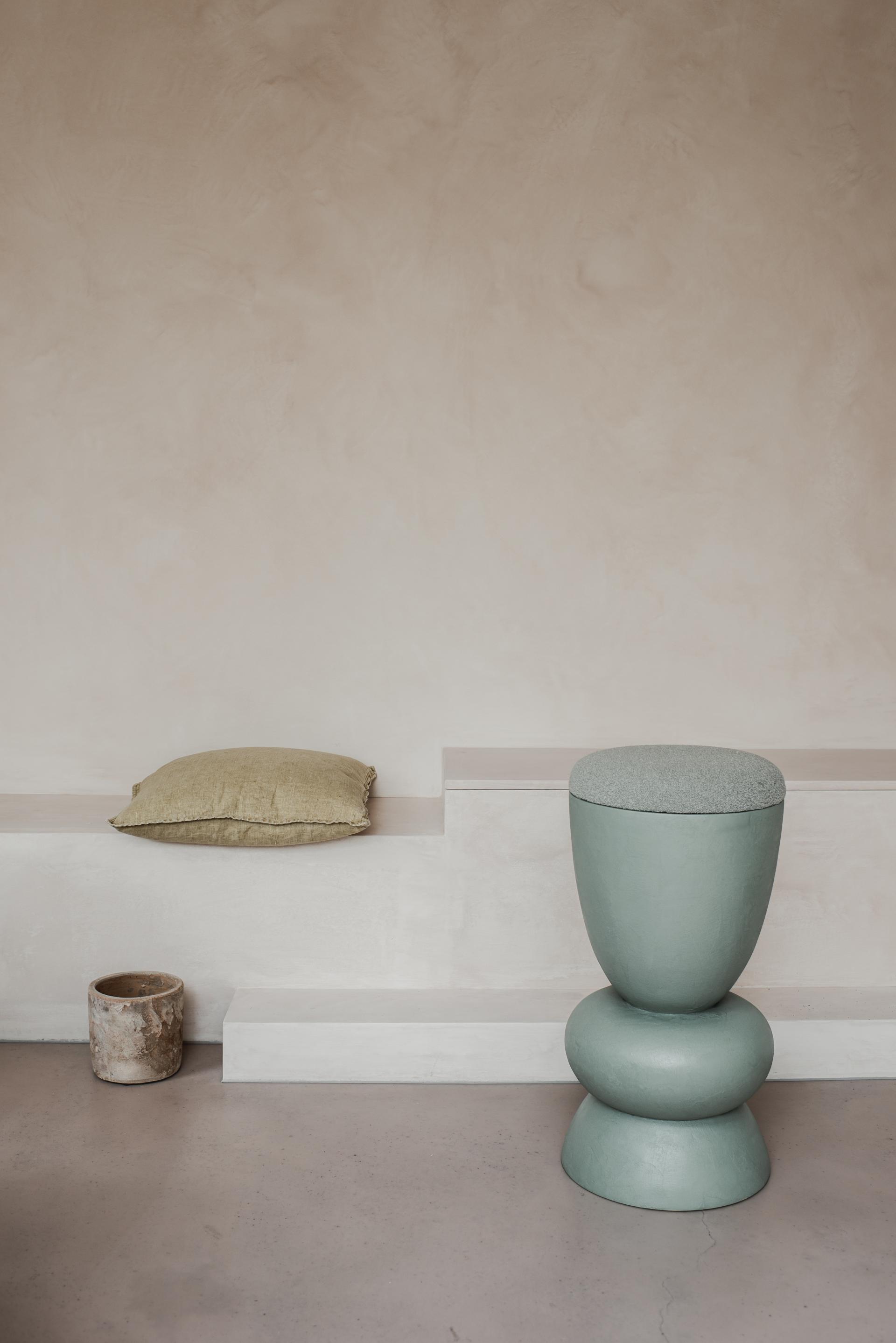 Curvaceous green stool with a textured top adjacent to a pillow on a minimalist indoor setting.