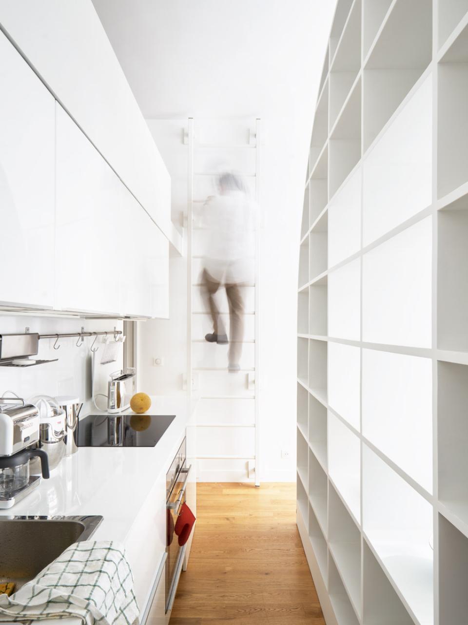 Elegant kitchen design in a Parisian home featuring sleek white cabinetry and a person climbing a modern ladder.