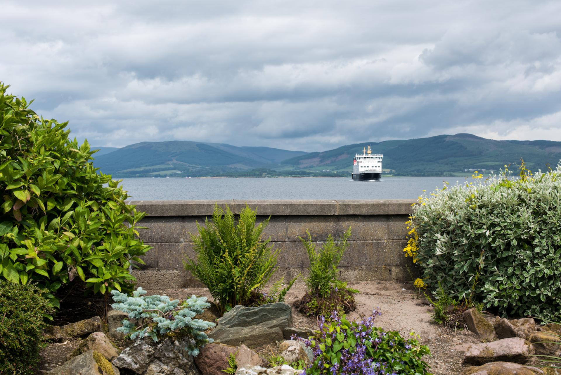 Scenic view of a ferry on water with lush greenery in the foreground and a mountainous backdrop.