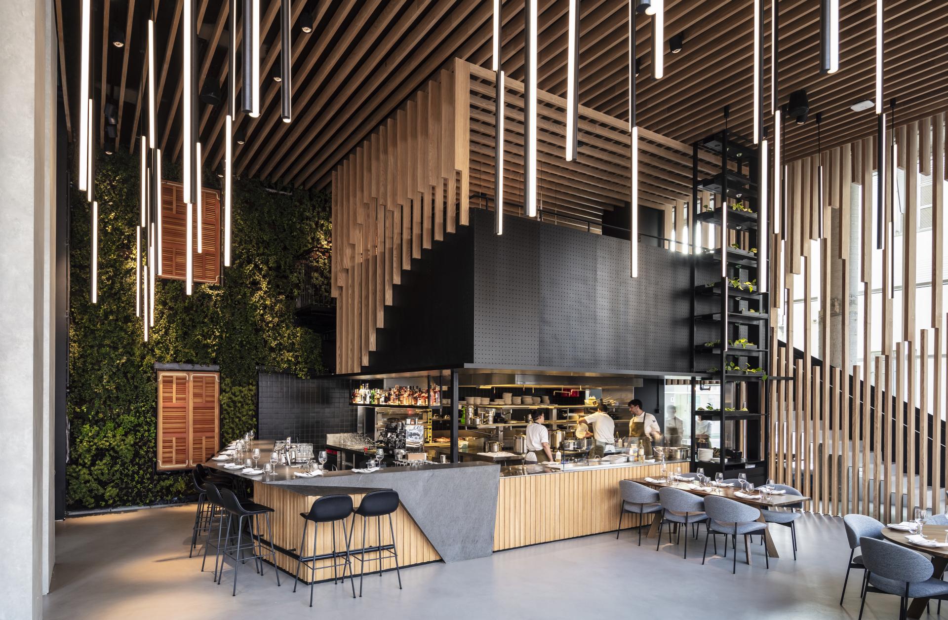 Modern restaurant interior featuring suspended wooden beams, lush greenery, and an open kitchen design by Kimmel Eshkolot Architects.