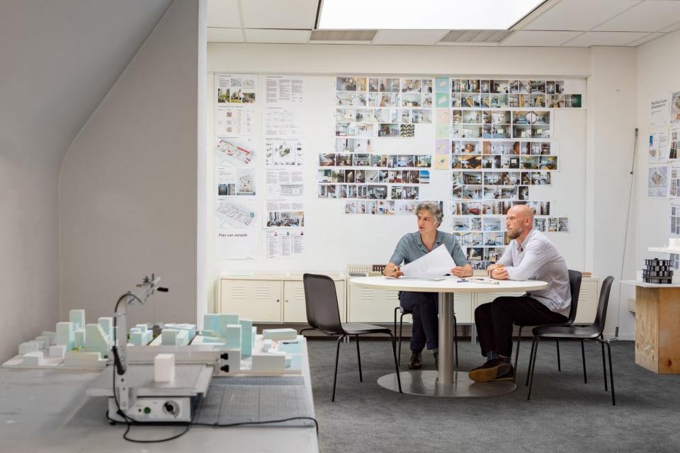 Two architects discussing plans at a round table, surrounded by design models and a wall filled with project photographs.