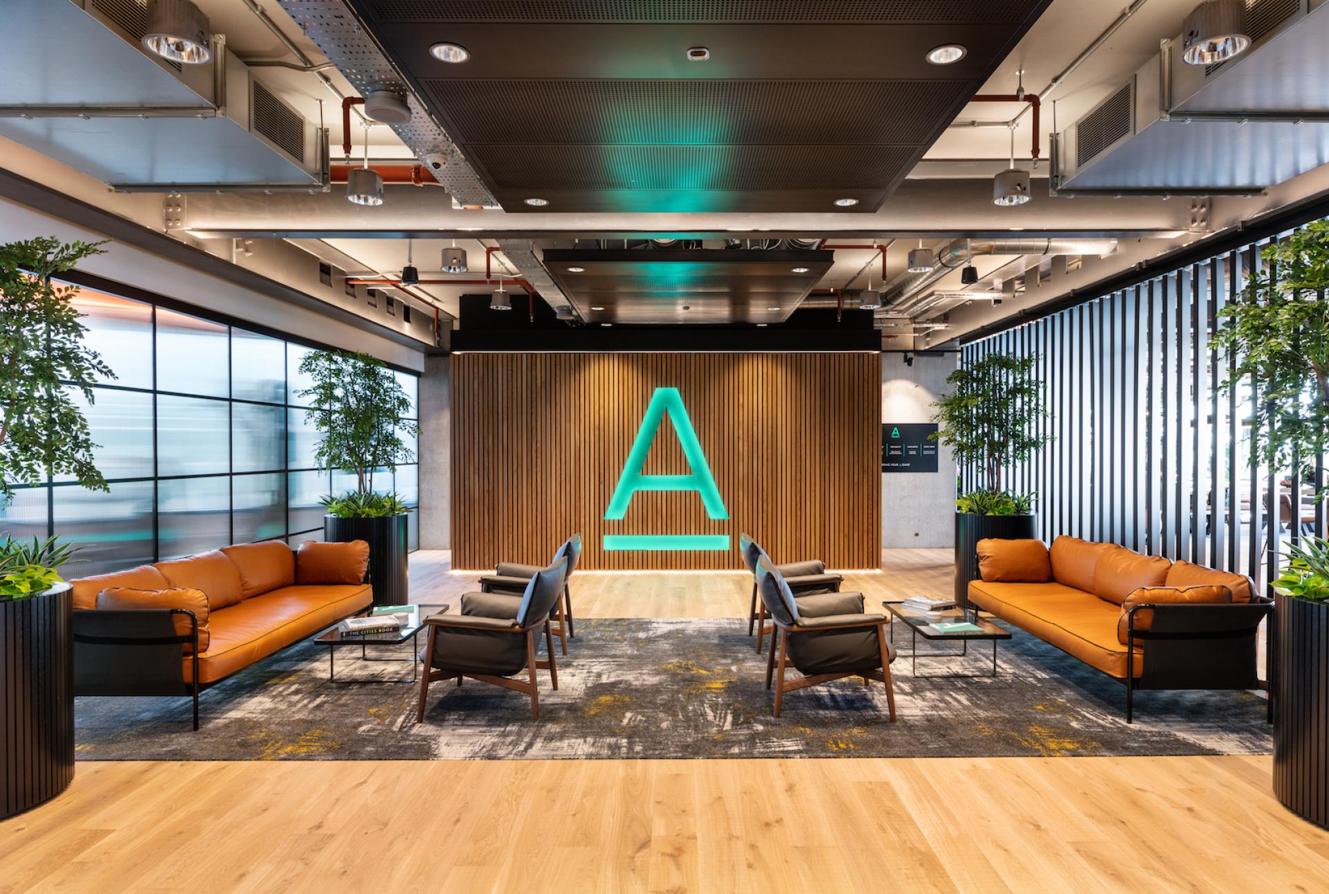 Modern workspace featuring sustainable design elements, comfortable seating, greenery, and an illuminated logo emphasizing creativity and collaboration.