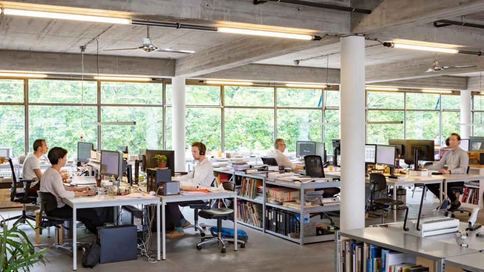 Brightly lit architects' studio featuring multiple workstations, plants, and large windows overlooking greenery.