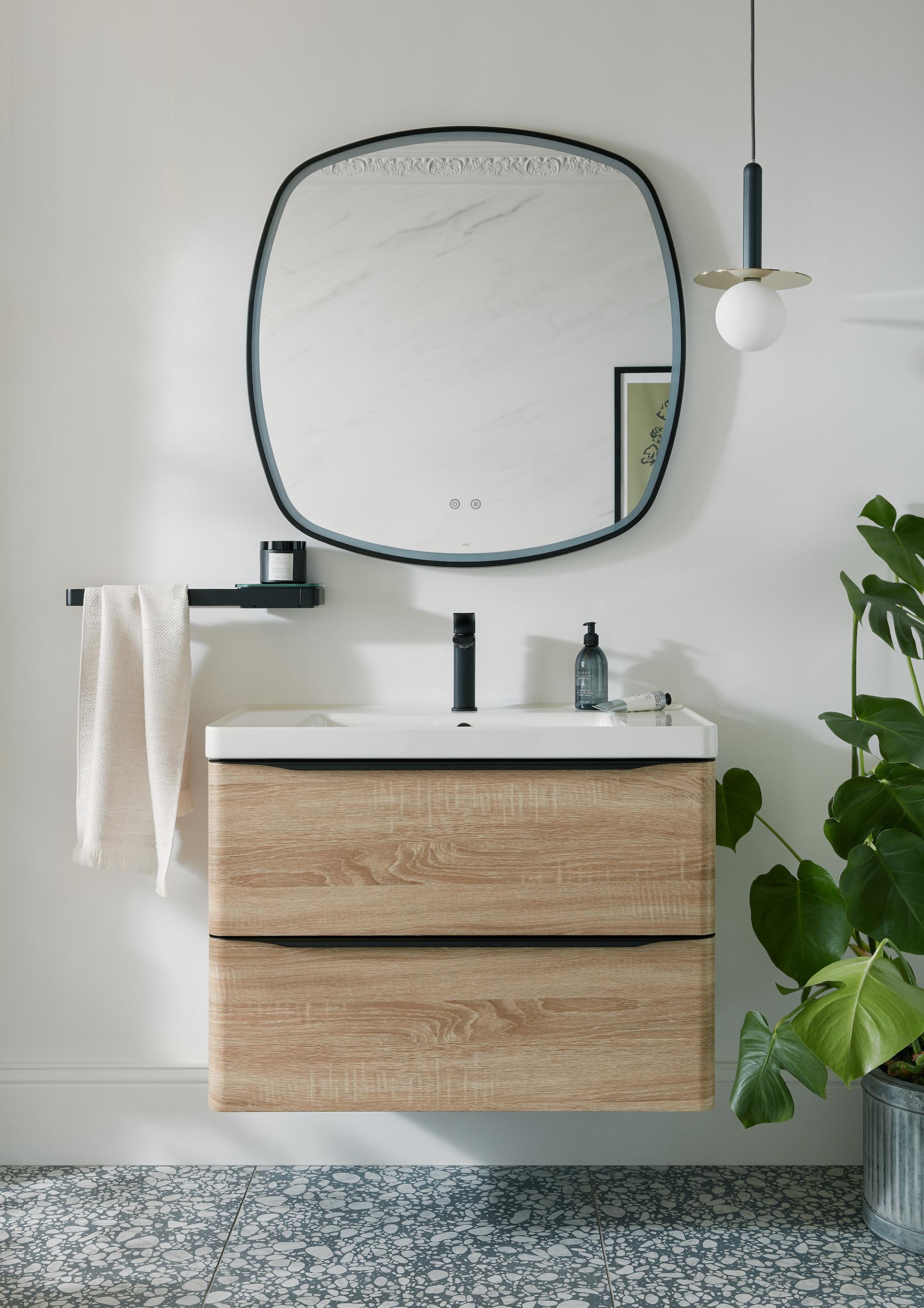 Modern bathroom featuring VADO's stylish cabinetry, round mirror, and elegant brassware, complemented by greenery and patterned flooring.