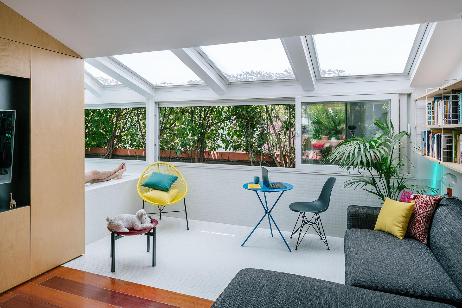 Oversized bathtub with a view in a sunlit attic, featuring modern furniture and greenery outside.
