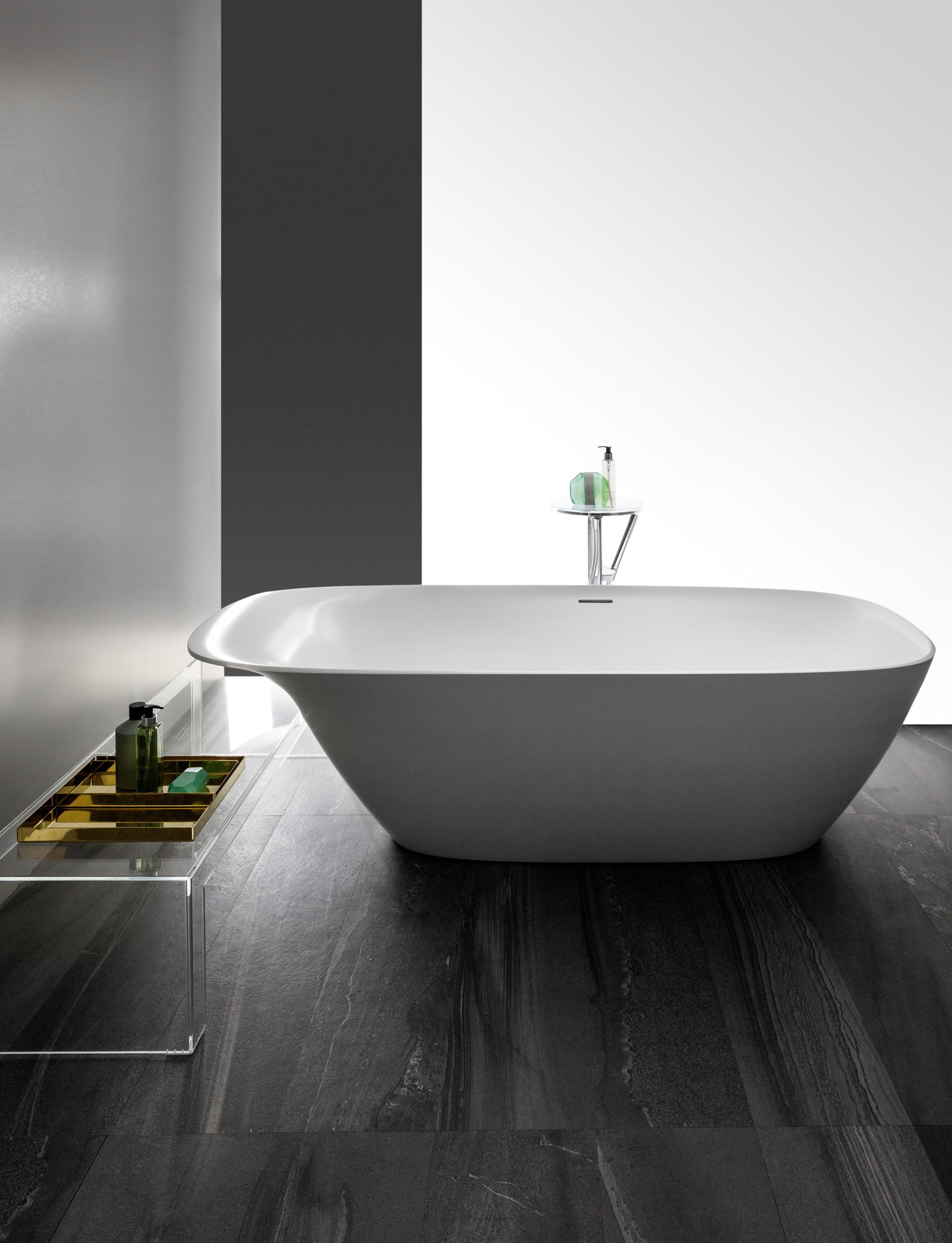 Modern freestanding bathtub by LAUFEN, accompanied by a minimalist side table with toiletries on a dark stone floor.
