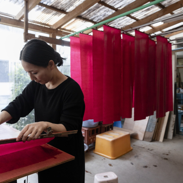 Artisan applying natural dye to fabric in a workshop, surrounded by vibrant red textiles hanging to dry.