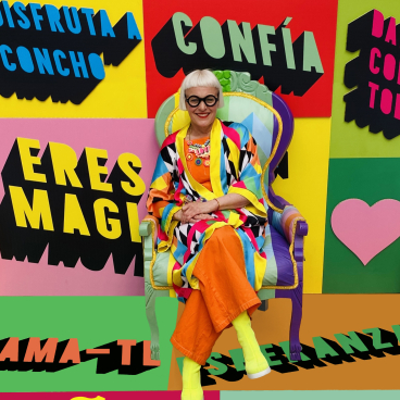 Creative figure in colorful outfit seated in vibrant chair against background of bold, motivational designs at Clerkenwell Design Week.