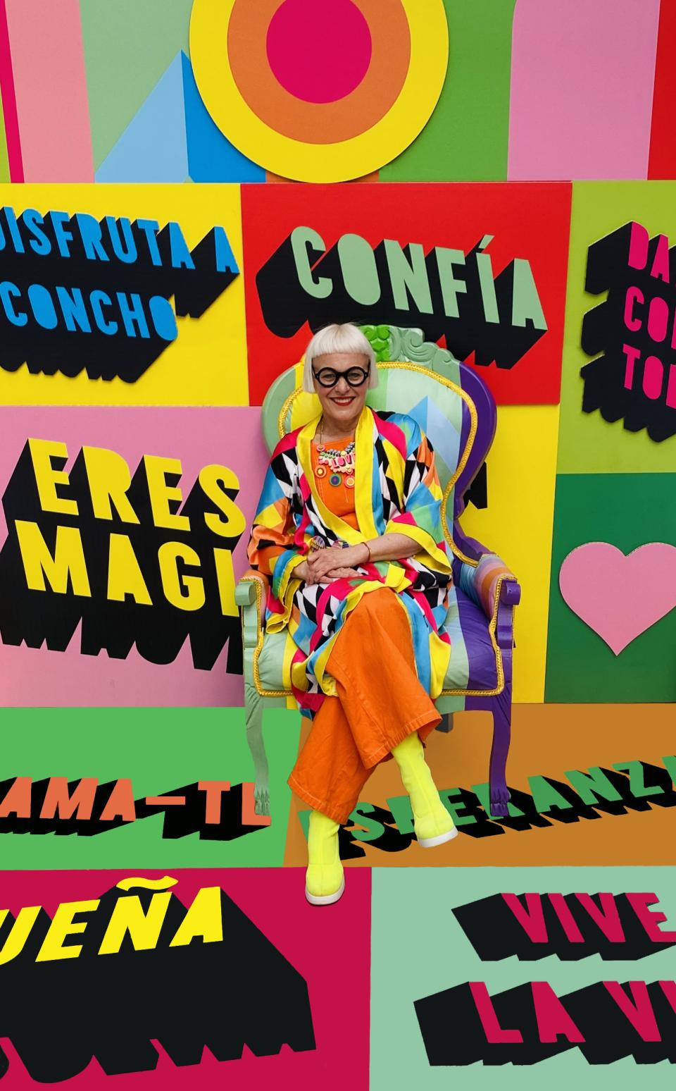 Creative figure in colorful outfit seated in vibrant chair against background of bold, motivational designs at Clerkenwell Design Week.