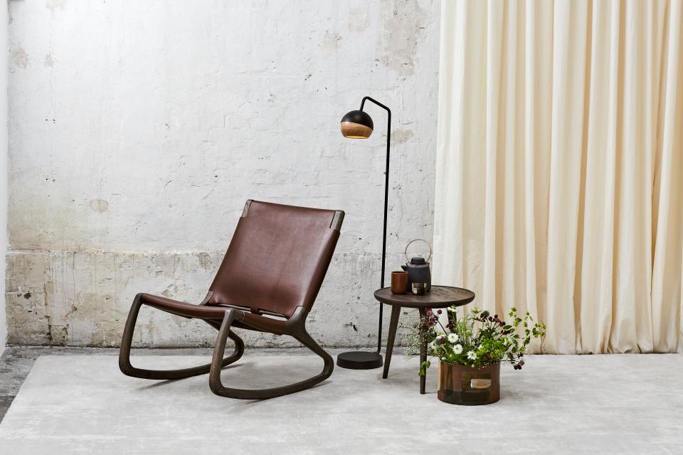 Elegant rocking chair by Mater with leather seating, accompanied by stylish lamp and table with flowers, against a textured wall.