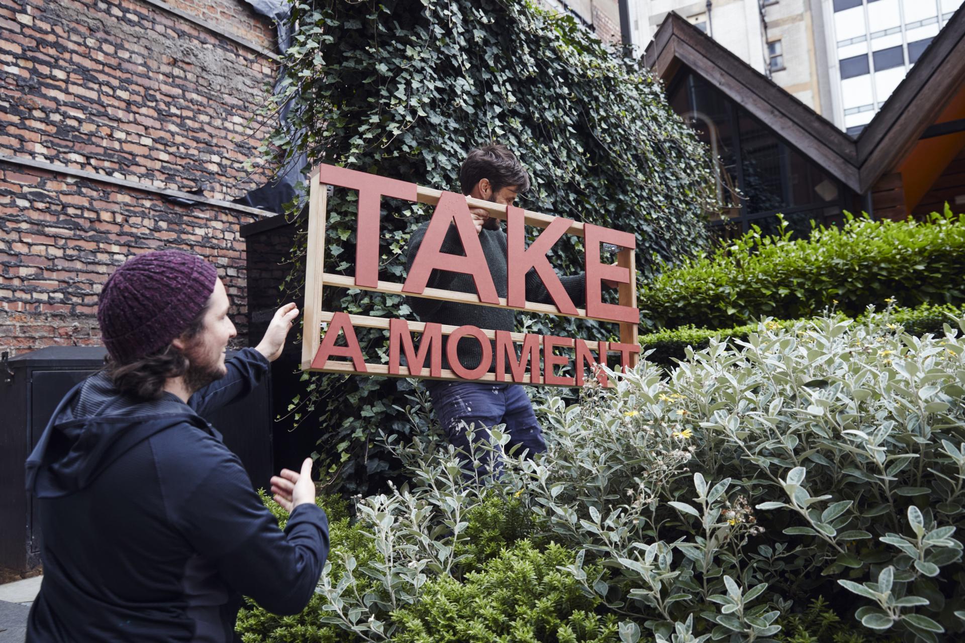 Two individuals installing a vibrant "Take A Moment" sign among greenery in Manchester's NOMA neighborhood.