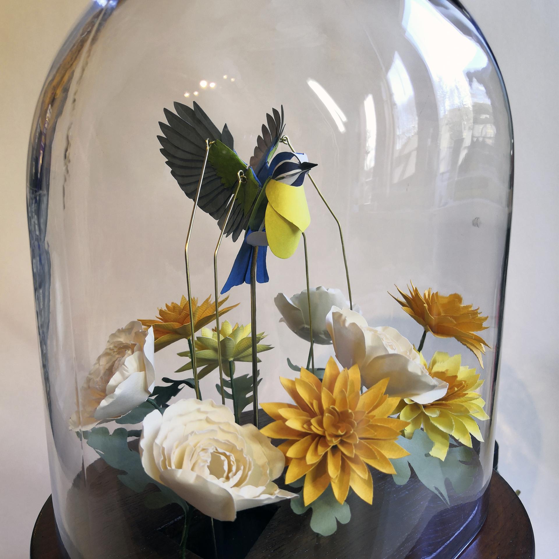 Colorful paper flowers and a bird sculpture are displayed under a glass dome at the Great Northern Contemporary Craft Fair.