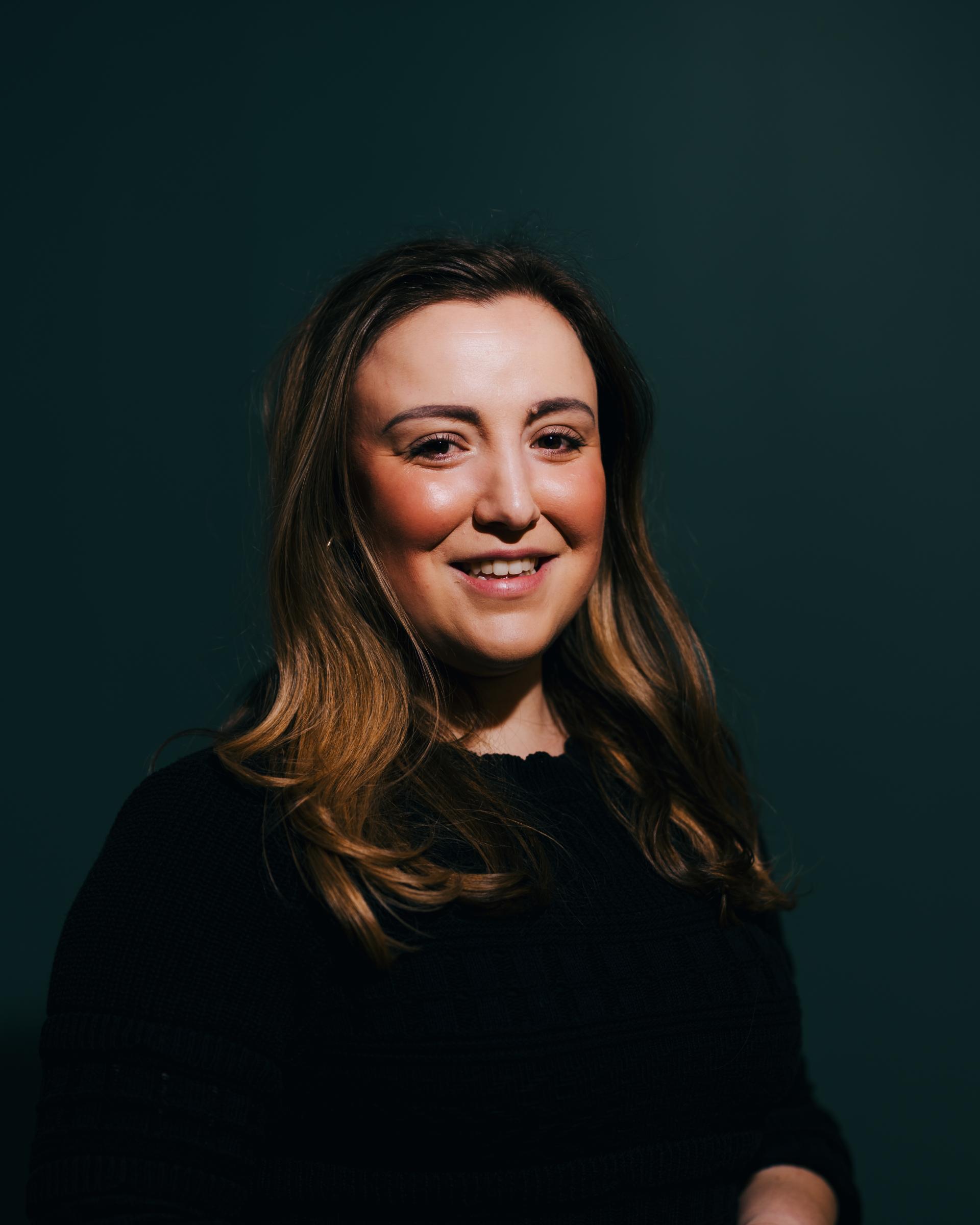 Smiling woman with long hair, wearing a black sweater, against a dark green background, discussing neurodiversity.