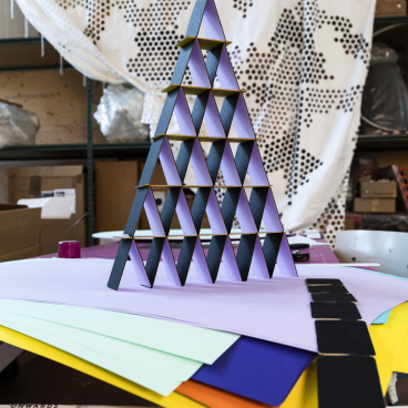 Colorful triangular paper structures arranged in a pyramid shape, with various sheets of colored paper in the foreground.