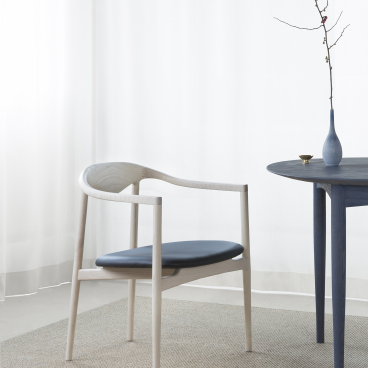 Contemporary dining chair in light wood with blue cushion, accompanied by a small round table and decorative vase.