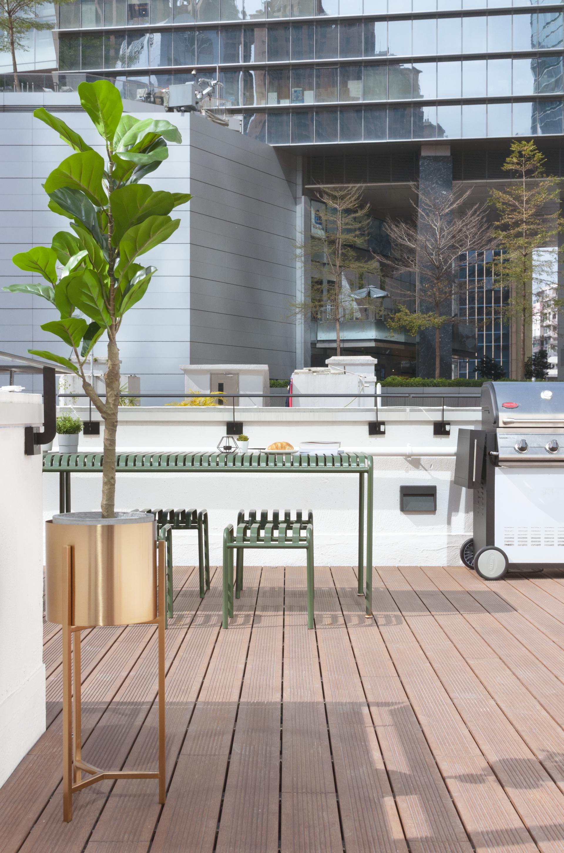 Contemporary outdoor workspace featuring a dining table, green chairs, potted plant, and barbecue grill in a revitalized urban setting.