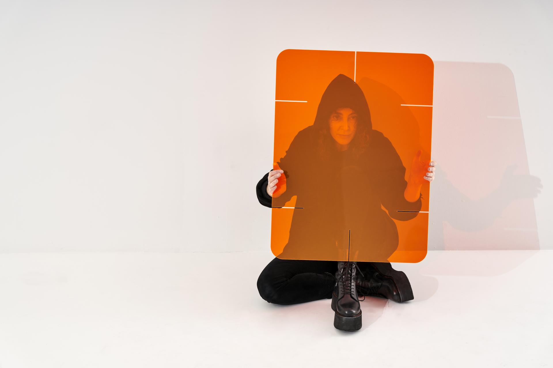 A person seated on a white floor holds an orange acrylic divider, showcasing innovative design for social distancing.