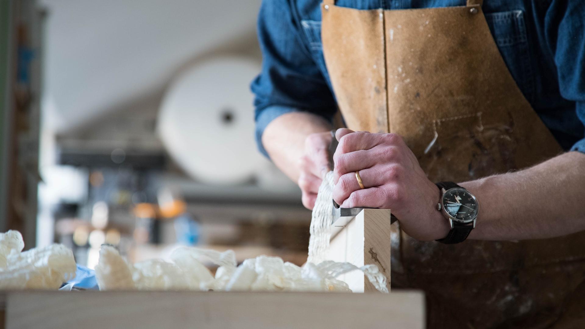 Craftsman shaping wood with a plane, surrounded by handmade furniture materials in a workshop that emphasizes local sourcing.