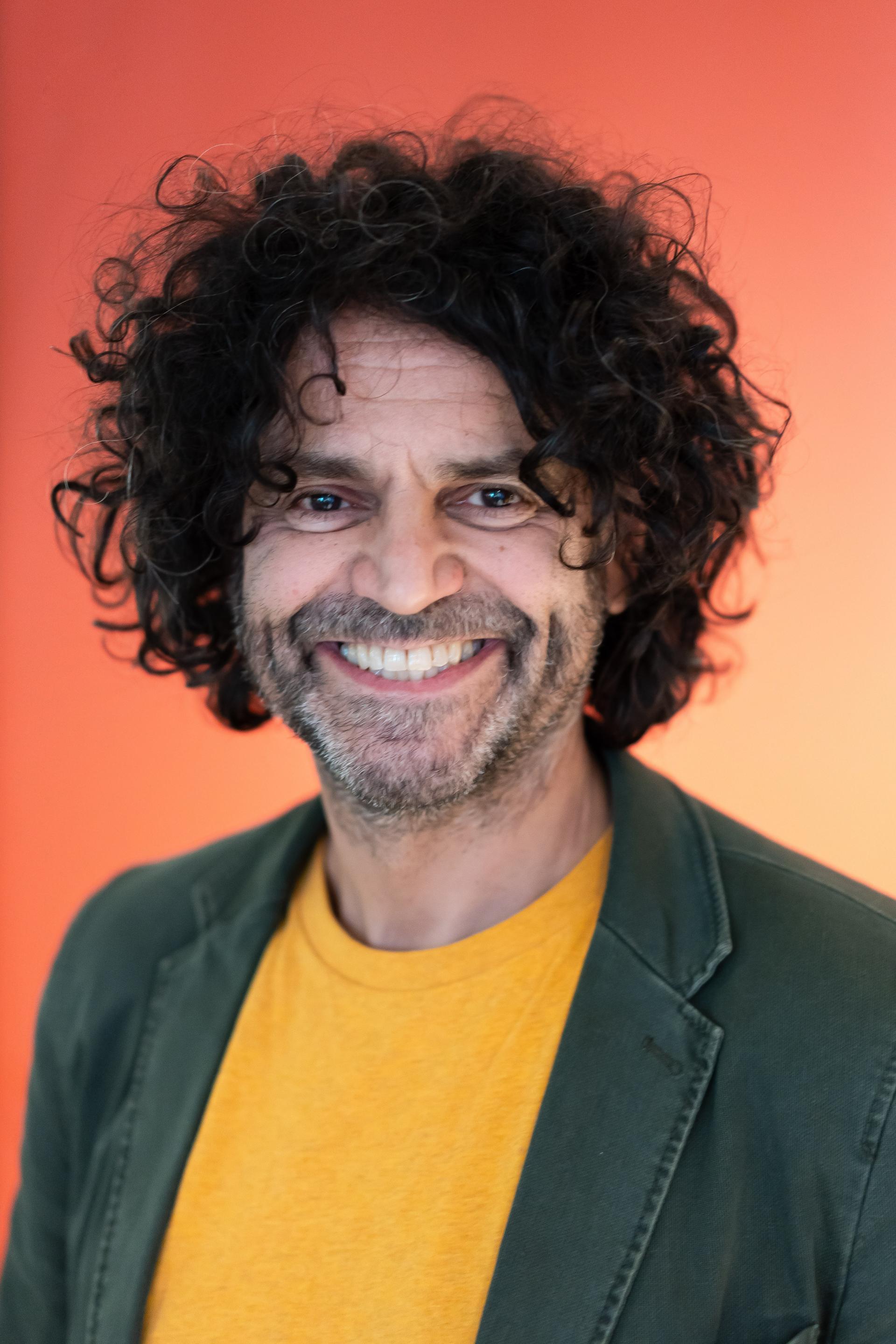 Smiling man with curly hair wearing a yellow shirt and green blazer, set against an orange background, representing workplace trends.