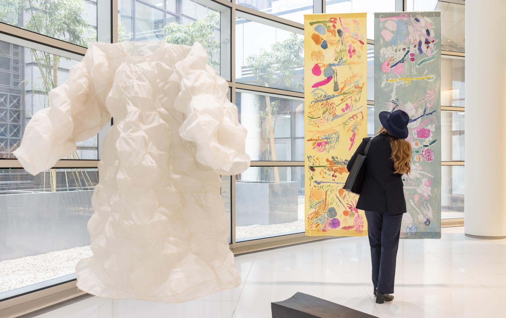 Sculptural ceramic dress installation with a viewer admiring colorful artwork in a contemporary art gallery setting.