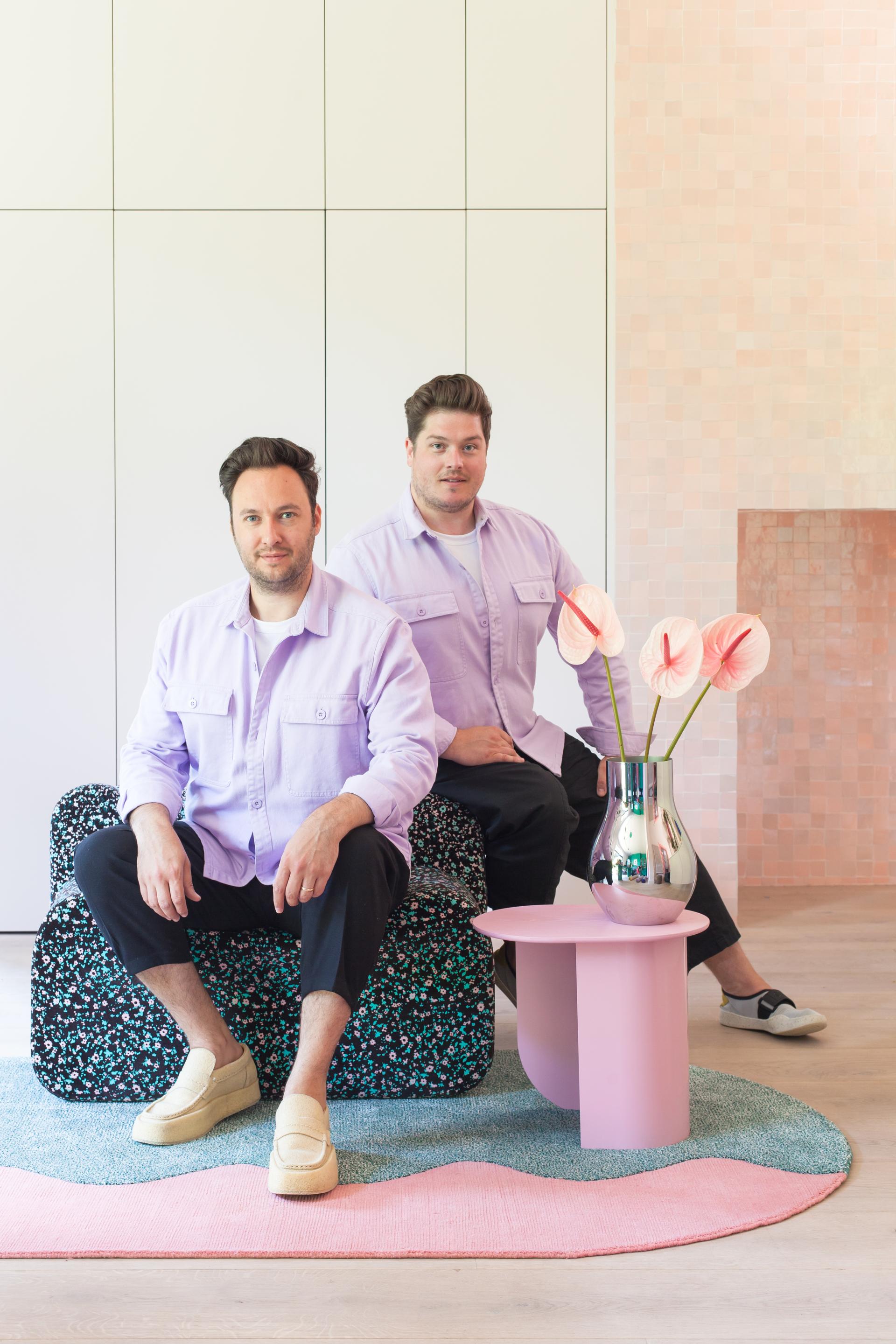 Design duo 2LG Studio seated on a patterned sofa, showcasing their FLOOR_STORY rug collection against an elegant interior backdrop.