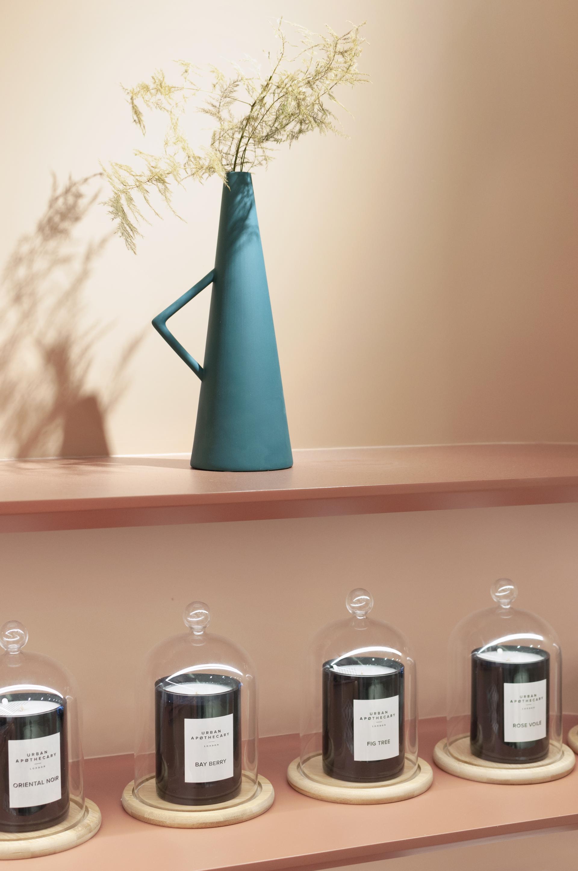 A teal sculptural vase holds dried foliage above a shelf featuring elegantly displayed candles in glass domes.
